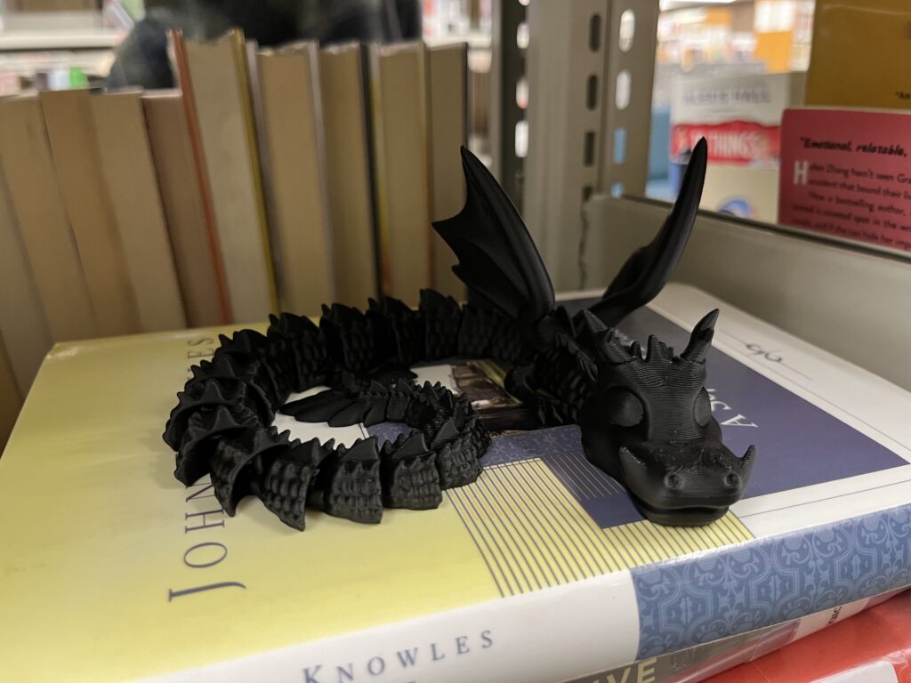A black flexi dragon curled up on top of a book in the stacks.