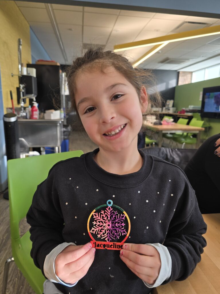 little girl smiling and holding a wooden snowflake ornament carved by our laser engraver. It's colored in and says her name. 